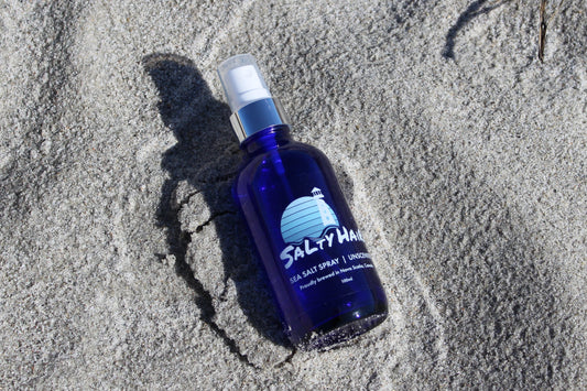 A blue glass bottle of unscented Salty Hair sea salt spray, sitting in sand at the beach.