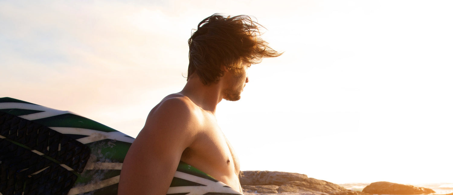 A male surfer stands on the beach, his saltwater-textured hair is windswept. He's holding his board and looking toward the ocean.