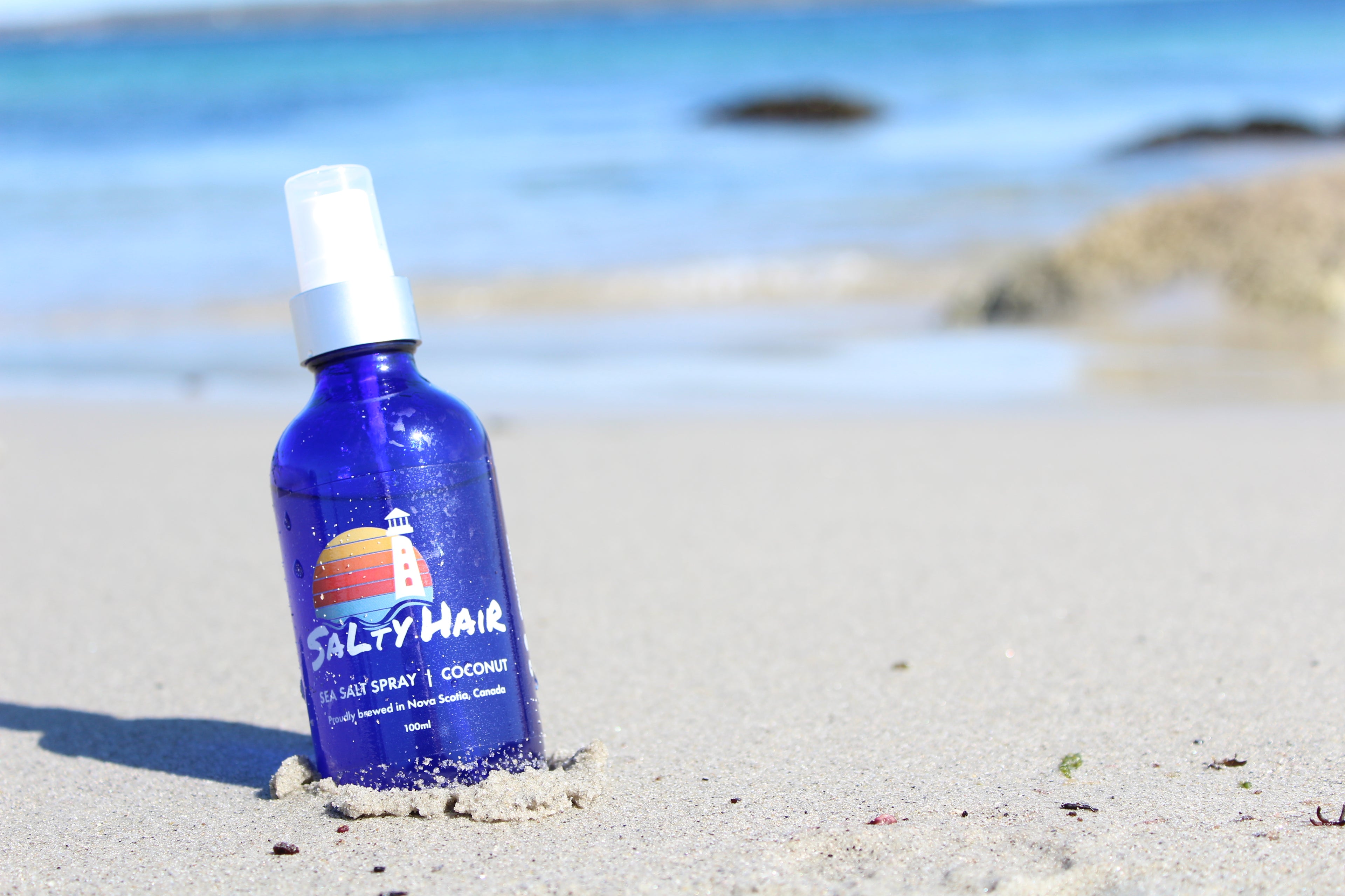 A blue glass bottle of Salty Hair sea salt spray in coconut scent is sitting on the sandy shoreline of a Nova Scotia beach.