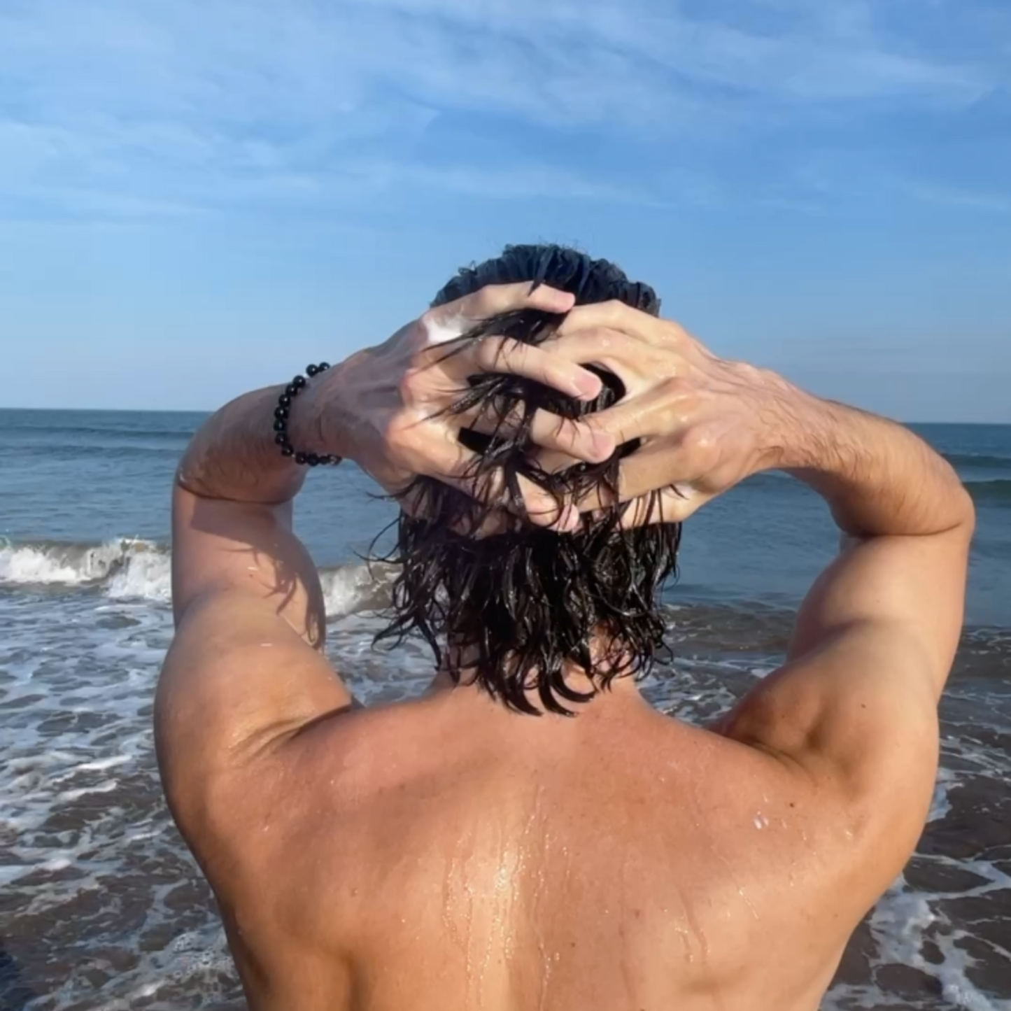 Person with wet hair on a beach washing their hair with Salty Hair Surf Shampoo, with ocean and sky in the background