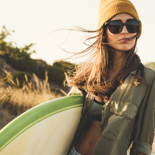 Woman with straight hair under a toque, carrying a surfboard, styled to tame flyaways, add volume, and refresh hair with Salty Hair Sea Salt Spray.